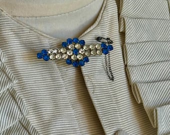 May include: A vintage brooch with a blue and clear rhinestone design. The brooch is attached to a cream-colored jacket with a safety pin and chain. The jacket has a textured, striped pattern.