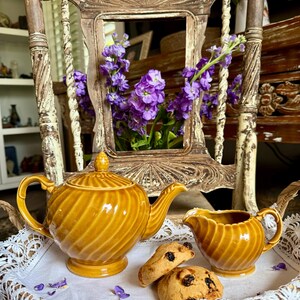 May include: A vintage tea set, including a teapot and creamer, in a warm yellow-brown hue, with a spiral design. Two cookies sit on a wicker tray. The background features purple flowers and a decorative chair.