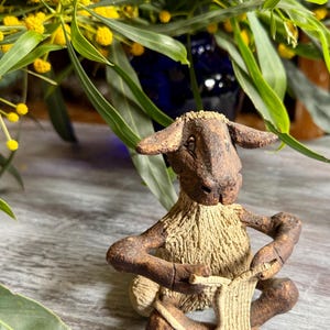 Puede incluir: Una figura de cerámica marrón y beige de una oveja sentada con las piernas cruzadas. La oveja tiene un cuerpo texturizado que parece lana y sostiene un pequeño trozo de material plano y rectangular en sus manos como si estuviera tejiendo.