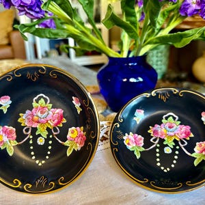 May include: Two black decorative plates with gold rims. Each plate features a floral design with pink, yellow, and blue roses. The plates are on a white lace tablecloth, with a blue vase and flowers in the background.