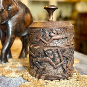 May include: A dark brown, carved wooden container with a lid and a handle. The container features detailed relief carvings of human figures. An intricately carved wooden elephant statue is in the background. The container sits on a woven mat.