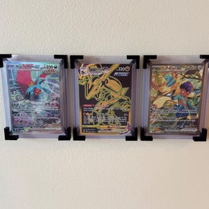 May include: Three framed Pokémon trading cards. The cards feature colorful illustrations of various Pokémon characters. Each card is encased in a clear protective sleeve and mounted with black corner supports.