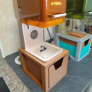 Puede incluir: Una impresora 3D de resina xTool con una cubierta protectora naranja translúcida con el nombre de la marca impreso en blanco. La máquina tiene una placa de construcción de metal plateado, un botón de parada de emergencia rojo y una base de almacenamiento marrón.