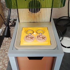 以下が含まれることがあります： グレーのフレームとオレンジの引き出しを備えた3Dプリンター。黄色のビルドプレート上に、金属製のリングが付いた2つの円形木製キーホルダーが置かれている。各キーホルダーには「FCR」という文字と、その上に小さなロゴが刻印されている。
