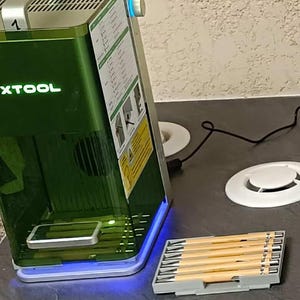 May include: A green and silver laser engraving machine with the word "xtool" on the front. The machine has a clear window and a blue light at the base. A tray of wooden tools sits in front of the machine.