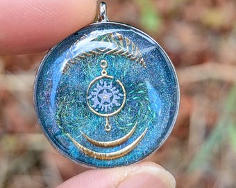 May include: A round pendant with a silver-colored bail. The pendant features a blue and green background with gold-colored celestial designs, including a sun, crescent moons, and a starburst. The pendant is held by a person's fingers.
