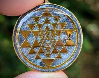May include: A gold-colored pendant with a Sri Yantra design. The pendant features a gold-colored outer rim and a gold-colored bail. The Sri Yantra design is composed of gold-colored triangles on a silver and gray background.