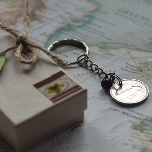 May include: A small brown box with a green heart-shaped tag tied with twine. The box is sitting on a map with a silver keychain with a coin attached. The coin is silver and has a 10p inscription.