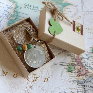 May include: A small brown box with a green heart on the lid. The box contains a silver key chain with a green and brown bead charm and a silver coin with a fish design and the text "10P".
