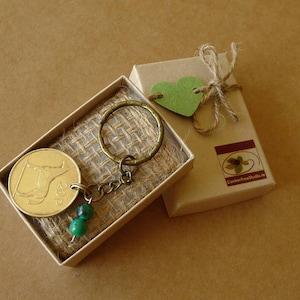 May include: A gold-colored coin keychain with a bull design, green beads, and a bronze-colored ring, presented in a small box with a burlap lining. A small gift box with a green heart tag and twine is next to it. The boxes are on a brown surface.