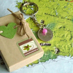 May include: A keychain with a silver coin pendant and a pink bead, presented in a small, brown gift box. The box is adorned with a green heart-shaped tag and a sticker with the text "ConkerTreeStudio.ie". The background is a map of Ireland.