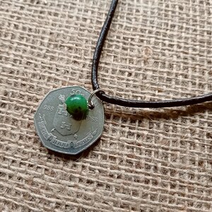 Op de afbeelding: Een groene glazen kraal hangt aan een bruin leren koord dat is bevestigd aan een zilveren Ierse munt van 50 pence. De munt is gedateerd 1988 en heeft de tekst "ÉIRE 1000 PENCE DUBLIN".