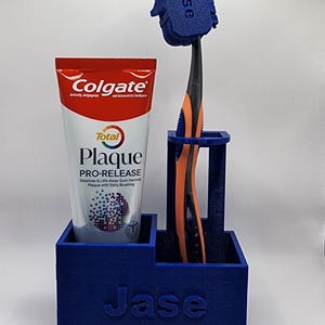 May include: A blue toothbrush holder with the name "Jase" on the front, holding a toothbrush and a tube of Colgate toothpaste. The toothbrush has a blue head and an orange and black handle. The toothpaste tube is white and red.