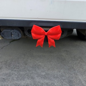 May include: A bright red velvet bow attached to the trailer hitch of a vehicle. The bow is centered, with the vehicle's bumper and hitch components visible in the background. The vehicle is light-colored, and the ground is concrete.