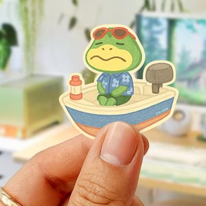 May include: A sticker featuring a cartoon frog in a boat, wearing sunglasses and a Hawaiian shirt. The boat is blue and beige, with a small red and white bottle. The frog has a sad expression.