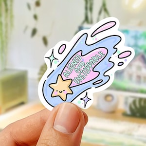 May include: A sticker with a cartoon star and the words "Aligned and Unstoppable" in a blue and pink design. The sticker has a white background and is held in a hand.