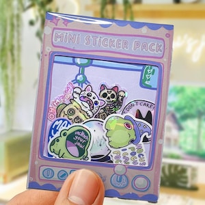 May include: A mini sticker pack with cartoon stickers, including phrases like "unlock your jaw" and "don't care". The pack is designed to resemble a claw machine, with a light purple and blue colour scheme.