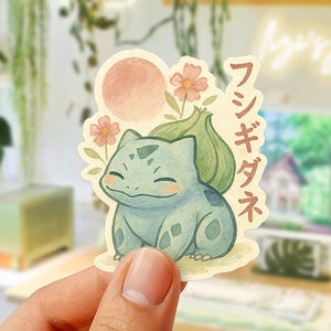 Puede incluir: Una pegatina con una ilustración de acuarela de un personaje de Bulbasaur con un sol rosa y flores. El texto japonés está impreso verticalmente en el lado derecho de la pegatina. La pegatina se sostiene en una mano.