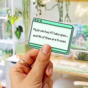 May include: A mint green sticker in the shape of a computer window, held by a hand. The sticker reads, "My brain has 47 tabs open... and 46 of them are frozen." The background includes a desk, plants, and a computer monitor.