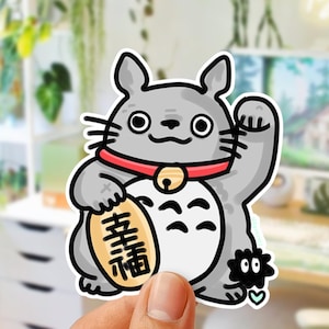 Könnte beinhalten: Ein grauer Totoro-inspirierter Aufkleber mit rotem Halsband und goldener Glocke. Der Aufkleber zeigt einen winkenden Arm und eine Goldmünze mit japanischen Schriftzeichen. Ein kleines schwarzes Wesen befindet sich neben dem Totoro. Der Aufkleber wird von einer Hand gehalten.