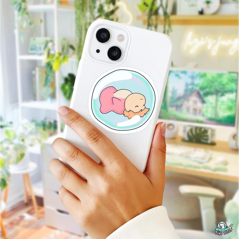 May include: White phone case with a circular sticker featuring a cartoon baby sleeping in a bubble. The baby is wearing a pink outfit and has light brown hair. The bubble is outlined in light blue.