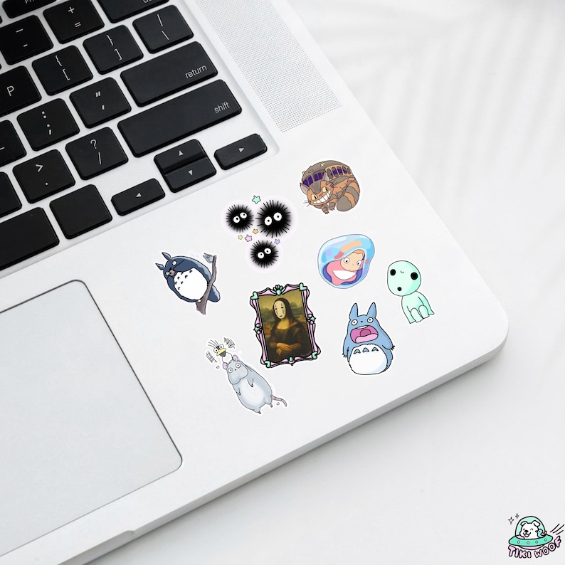 May include: A selection of colourful stickers featuring cartoon characters and artwork. The stickers include images of Totoro, the Mona Lisa, and other whimsical designs. The stickers are placed on a white laptop.