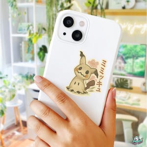 K&ouml;nnte beinhalten: Wei&szlig;es Handycase mit einem Mimikyu-Aufkleber. Der Aufkleber zeigt ein Cartoon-Pok&eacute;mon vom Typ Geist mit beigem K&ouml;rper, gezacktem Mund und dunkelbraunem Schwanz. Rosa Blumenakzente und japanischer Text sind ebenfalls vorhanden.