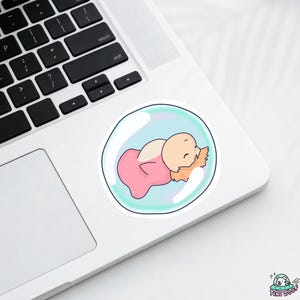 May include: A sticker featuring a cartoon baby sleeping inside a bubble. The baby is wearing a pink outfit and has light orange hair. The bubble has a light blue interior and a clear outline. The sticker is on a white surface.