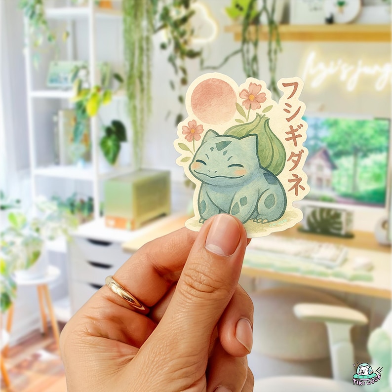 May include: A sticker featuring a watercolor illustration of a Bulbasaur character with a pink sun and flowers. Japanese text is printed vertically on the right side of the sticker. The sticker is held in a hand.