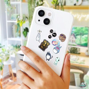 May include: A white smartphone adorned with various stickers of cartoon characters and art. The stickers include a depiction of the Mona Lisa, a blue Totoro, and other whimsical designs. The phone is held in a person's hand.
