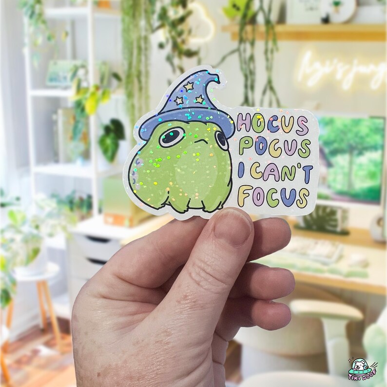 May include: A holographic sticker featuring a green frog wearing a blue wizard hat with stars. The sticker reads "Hocus Pocus I Can't Focus" in colorful bubble letters. The frog has a grumpy expression.