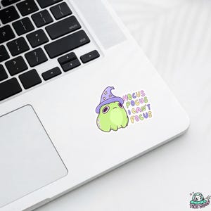 May include: A sticker featuring a cartoon frog wearing a purple wizard hat. The frog is light green with a grumpy expression. Text on the sticker reads "Hocus Pocus I Can't Focus" in pastel colors. The sticker is on a white laptop.