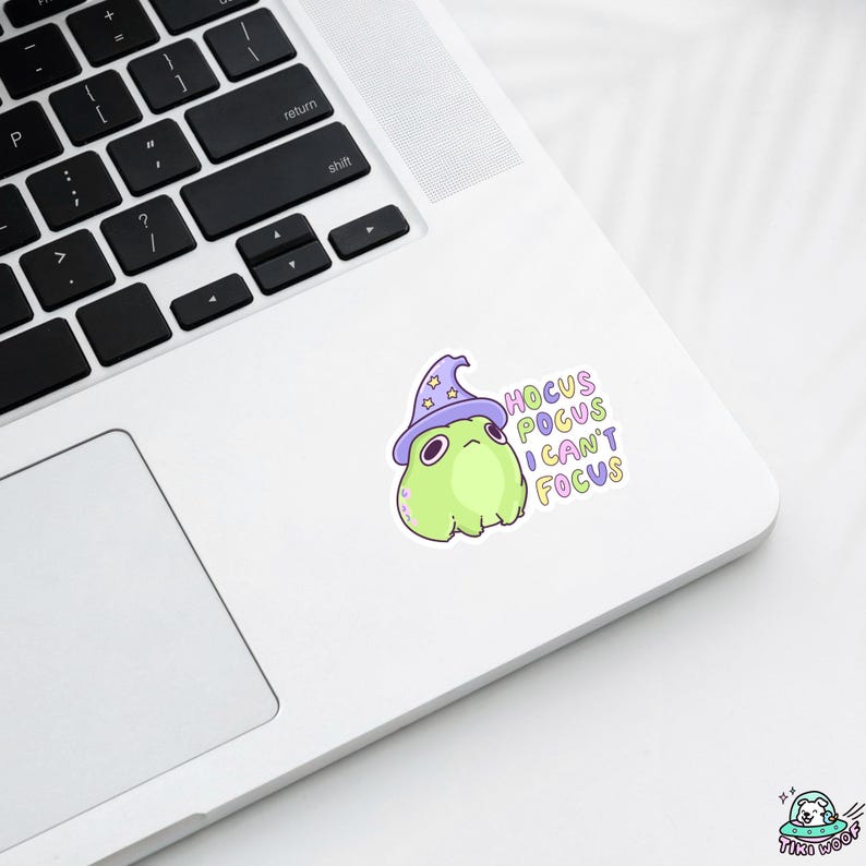 May include: A sticker featuring a cartoon frog wearing a purple wizard hat. The frog is light green with a grumpy expression. Text on the sticker reads "Hocus Pocus I Can't Focus" in pastel colors. The sticker is on a white laptop.