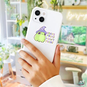 May include: A white smartphone with a sticker featuring a green cartoon creature wearing a purple witch hat. The sticker reads "Hocus Pocus I Can't Focus" in a playful font. The phone is held in a person's hand.