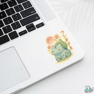 May include: A sticker featuring a watercolor illustration of a Bulbasaur character with Japanese text. The sticker has a light blue Bulbasaur, a peach-colored sun, and orange flowers. The sticker is on a white laptop.