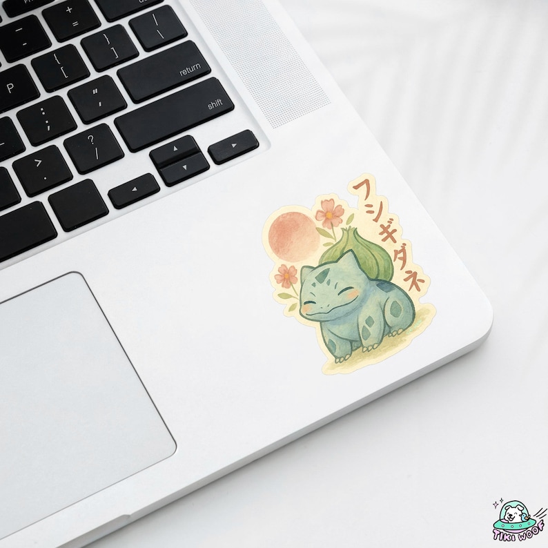 May include: A sticker featuring a watercolor illustration of a Bulbasaur character with Japanese text. The sticker has a light blue Bulbasaur, a peach-colored sun, and orange flowers. The sticker is on a white laptop.