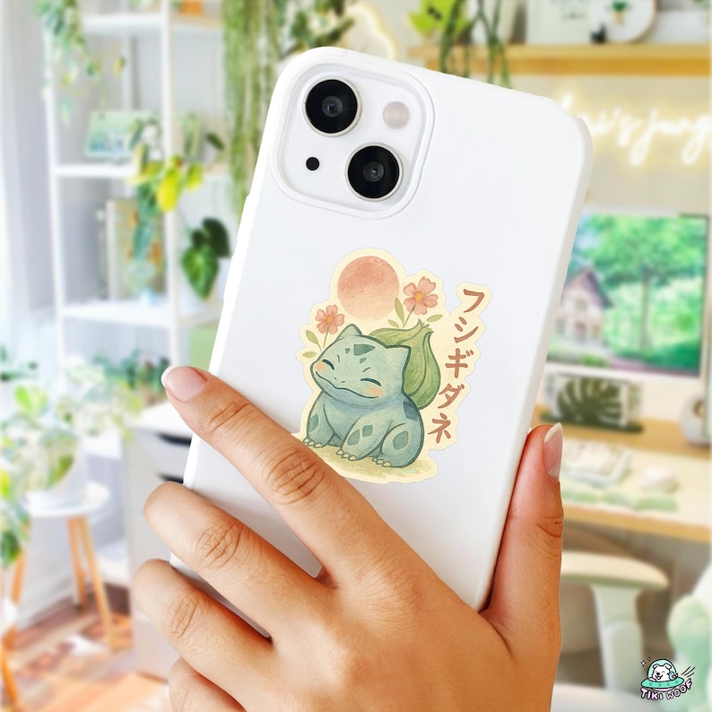May include: White iPhone case with a sticker featuring a cartoon Bulbasaur character, a sun, and flowers. Japanese text is also present. The sticker has a watercolor style with green, orange, and yellow colors.