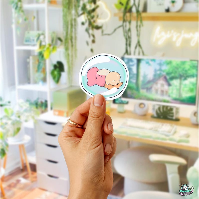 May include: A sticker featuring a cartoon baby in a bubble. The baby is depicted in pink and peach tones, with a light blue bubble outline. The sticker is held up in front of a blurred background of a desk and plants.