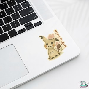 K&ouml;nnte beinhalten: Ein Aufkleber mit einem Cartoon-Mimikyu-Charakter mit einem l&auml;chelnden Gesicht, braunen Ohren und einem kleinen Schwanz. Der Aufkleber hat japanischen Text und rosa Blumenakzente. Der Aufkleber befindet sich auf einem wei&szlig;en Laptop.
