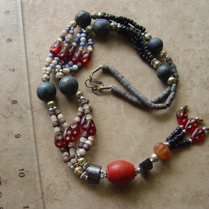 May include: A multi-strand necklace featuring a variety of beads in shades of red, black, gray, and cream. The necklace includes large, dark gray spherical beads, small red glass beads, and metallic accents. A ruler is in the background.