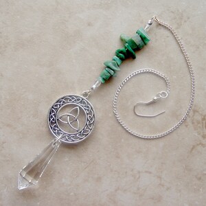 May include: A silver-toned pendulum with a clear crystal point and a Celtic knot design. The pendulum features a chain and is adorned with green gemstone chips. The design is suspended from a chain with a hook.