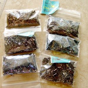 May include: Six small plastic bags filled with small, metallic-colored beads. The beads are primarily brown, with hints of gold and other colors. A ruler is visible in the upper left corner, and a small tag is visible in the upper right corner.