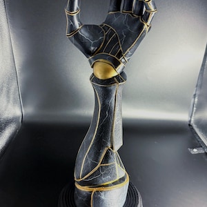 May include: A black and gold robotic hand sculpture. The hand is posed in a gesture, with intricate detailing and gold accents. The sculpture is mounted on a black base, creating a striking contrast. The overall design has a futuristic aesthetic.
