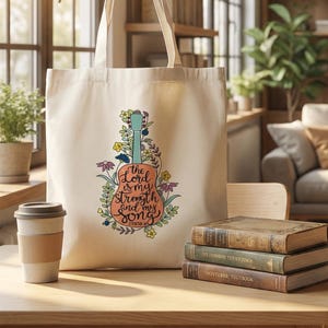 May include: Cream-colored canvas tote bag with a guitar design and the text "The Lord is my strength and my song." The bag has a roomy interior and a spacious design. The bag is durable and fits all essentials.