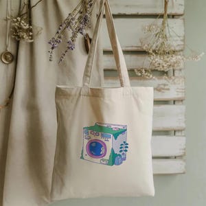 May include: Cream-colored tote bag featuring a whimsical illustration of a washing machine covered in greenery. The washing machine has a blue porthole, pink accents, and a green top. The bag has long handles for carrying.