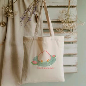May include: Beige canvas tote bag featuring a cartoon duck with its rear end in a pond, surrounded by lily pads. The text "i don't give a duck" is printed below the illustration. The bag has two long handles.