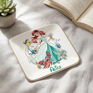 May include: Square ceramic jewelry dish with a gold rim featuring a colorful illustration of Ariel from The Little Mermaid. The dish has a gold necklace and ring on it, and the name "Alice" is printed on the bottom.