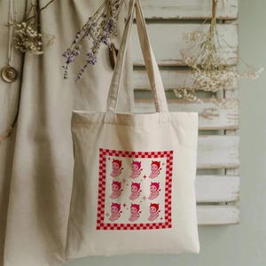Może przedstawiać: Kremowa torba typu tote z czerwono-białą kratką. Torba ma powtarzający się wzór różowych postaci diabła z rogami i ogonami. Torba wisi na neutralnym tle z suszonymi kwiatami.