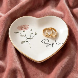 May include: Heart-shaped white ceramic ring dish with a gold rim, featuring a pink rose illustration and the name "Sophia" in script. Two gold rings rest on the dish, set against a pink velvet background.