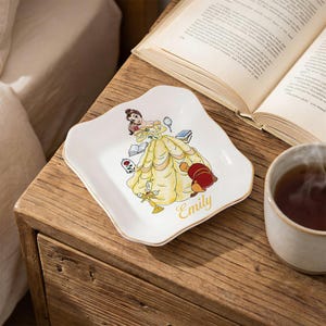 Könnte beinhalten: Eine weiße Schmuckschale mit gewelltem Rand und Goldrand, die eine Cartoon-Illustration einer Prinzessin in einem gelben Kleid zeigt. Der Name "Emily" ist in goldener Schrift geschrieben. Ein Buch und eine Teetasse sind im Hintergrund.